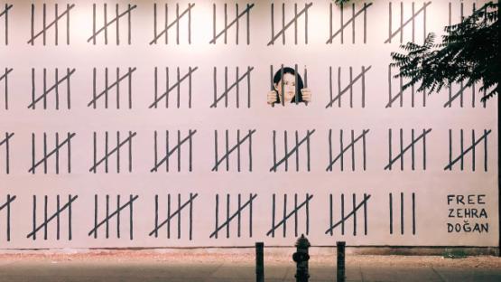 Tally marks painted on a wall, behind one set is a woman painted as though looking through jail bars, in the corner text reads 'Free Zehra Doğan'