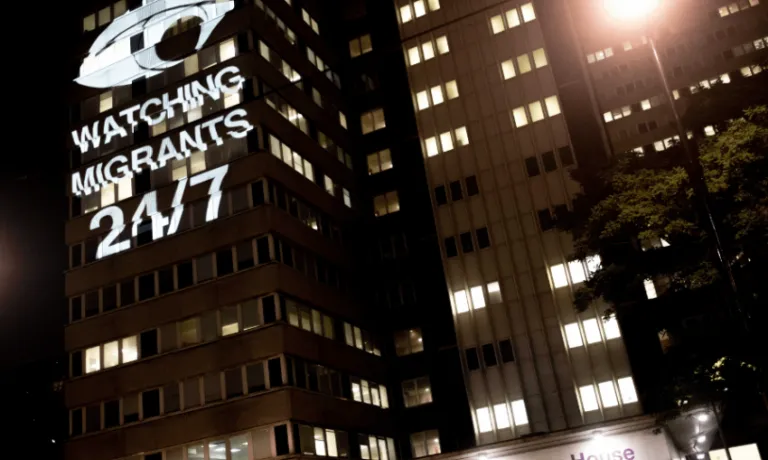 Office building at night with light projection showing watchful eye and reading "WATCHING MIGRANTS 24/7"