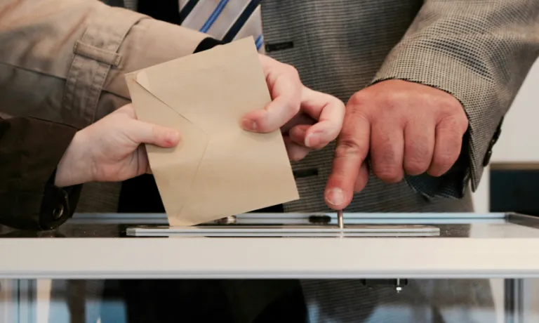 Image of three hands voting, casting a ballot paper into a box together.