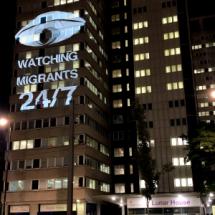 Light projection on UK Home Office building reading "WATCHING MIGRANTS 24/7"