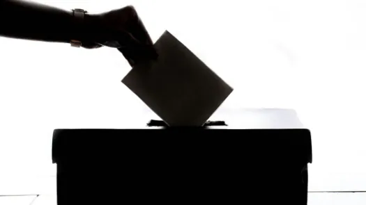 Black and white photograph depicting silhouette of hand inserting voting ballot into box.