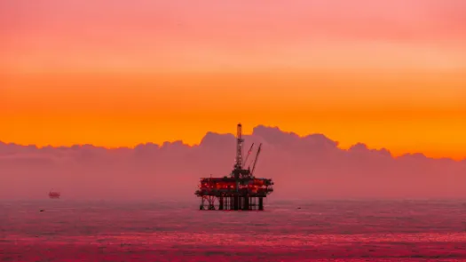 An offshore oil platform stands in the sea at sunset, framed by orange and pink skies, evoking the role of fossil fuel industries in debates about public subsidy, risk, and long-term harm.