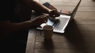 Manos en el teclado de un portátil junto a una taza de café, la pantalla está oculta
